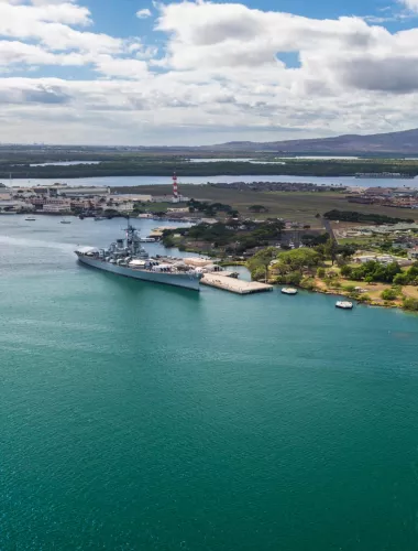 An aerial view of Pearl Harbor, featuring a historic battleship docked near lush greenery and calm blue waters.