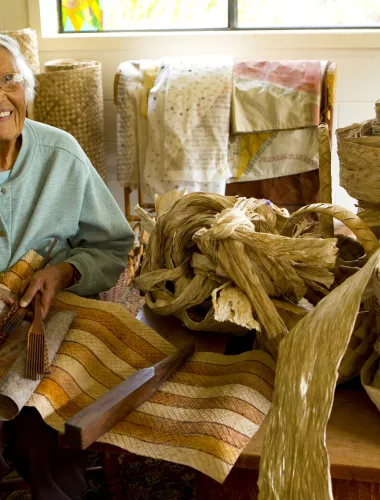 Elderly woman weaving traditional Hawaiian handicrafts with dried leaves in a cozy, light-filled room.