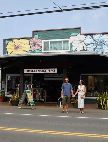Honokaa Couple holding hands outside the Honokaa Marketplace, a colorful building decorated with hibiscus murals, on a sunny day.