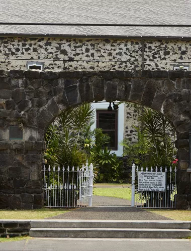 Stone building and white gate in Historic Kailua Village, surrounded by tropical greenery.