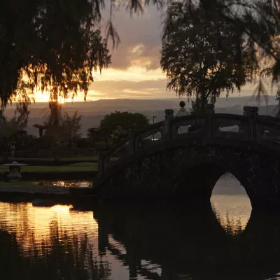 A scenic view of Liliuokalani Gardens, a Japanese garden, at sunset with a traditional arched bridge over a calm pond.