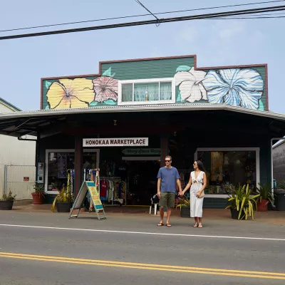 Couple holding hands outside the Honokaa Marketplace, a colorful building decorated with hibiscus murals, on a sunny day.