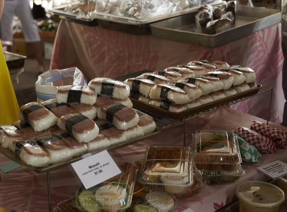 Tray of spam musubi and containers of butter mochi on display at a market stall, showcasing popular Hawaiian snacks and sweets.