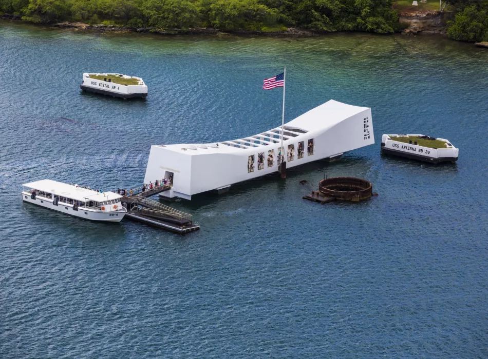 The Pearl Harbor USS Arizona Memorial, a white structure floating over the sunken battleship in calm waters, with nearby boats.